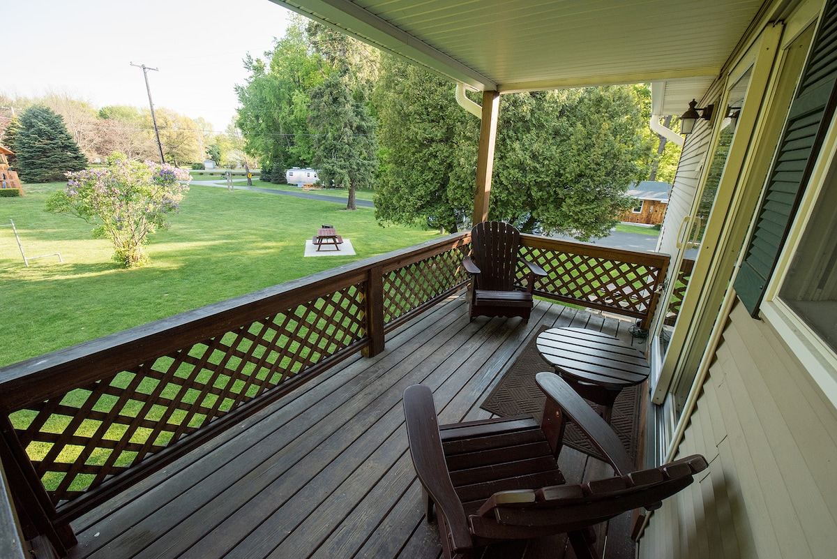 A covered porch is presented with two wooden chairs and a small table, surrounded by a wooden railing. Green grass and trees create a serene outdoor view, enhancing the connection to nature.