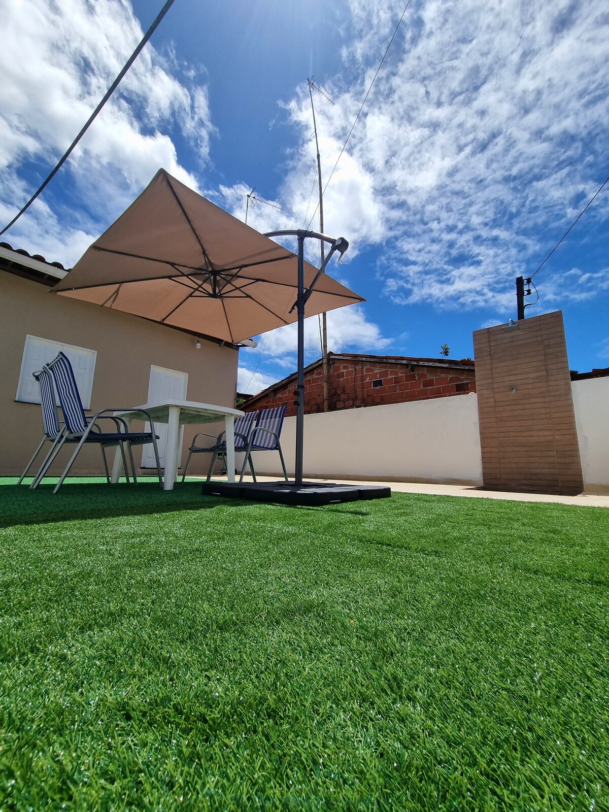 A spacious outdoor area features synthetic grass, providing a comfortable setting. A large umbrella offers shade over a dining table with four chairs, ideal for gatherings. The blue sky and scattered clouds complement the scene, creating a relaxing atmosphere for outdoor activities.
