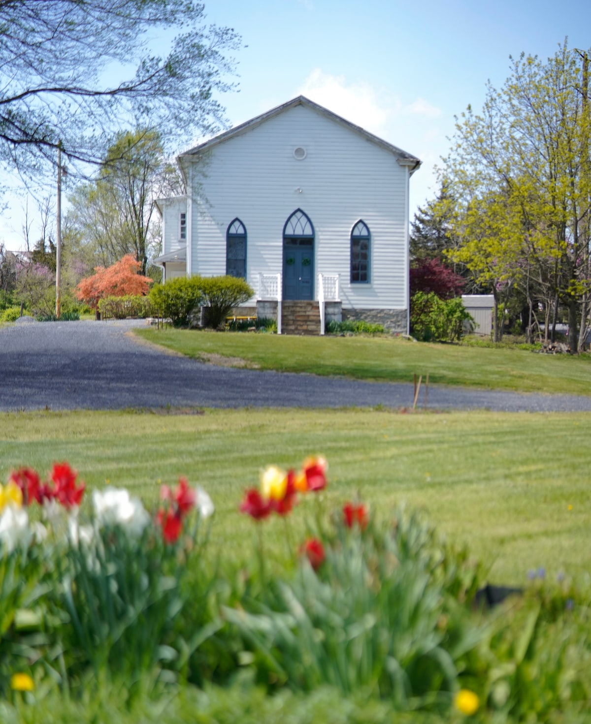 A charming white church building is set against a clear blue sky, surrounded by vibrant green grass and colorful flower beds featuring blooming tulips. The entrance is characterized by a large arched door flanked by decorative windows.