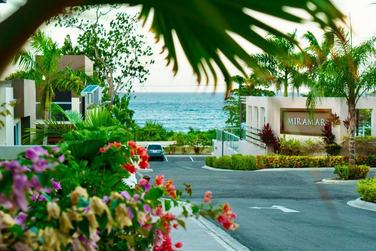 A gated entrance is framed by lush tropical plants and vibrant flowers, leading to the Miramar complex. The tranquil sea is visible beyond the buildings, with clear blue waters contrasting against the greenery. A sense of calm is present throughout the landscape.
