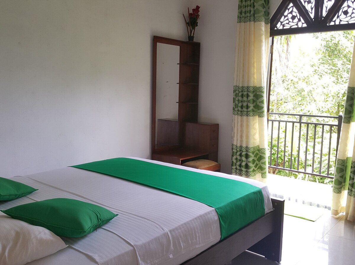 A bright private room features a spacious bed adorned with green accents. A dressing table is visible beside the bed, while large windows with sheer curtains open to a balcony. Lush greenery can be seen through the windows, enhancing the serene atmosphere.