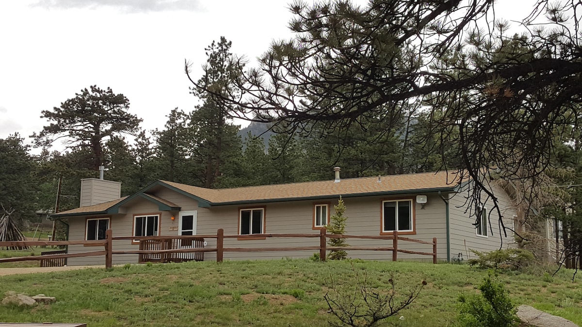 A spacious cabin is set against a backdrop of towering trees and mountains. Large windows and a welcoming porch are visible. The exterior is clad in neutral tones, complemented by a muted roof, creating a harmonious blend with the forested surroundings.