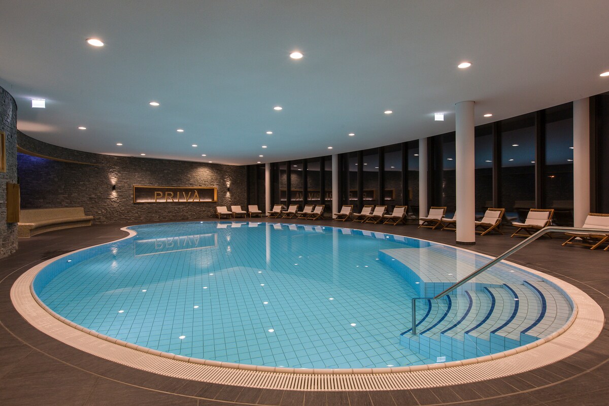 An indoor pool area is highlighted by smooth blue tiles and a gently curved shape. Several lounge chairs are arranged along the perimeter, providing ample relaxation space. Large windows create a bright atmosphere, allowing natural light to enhance the tranquil setting.