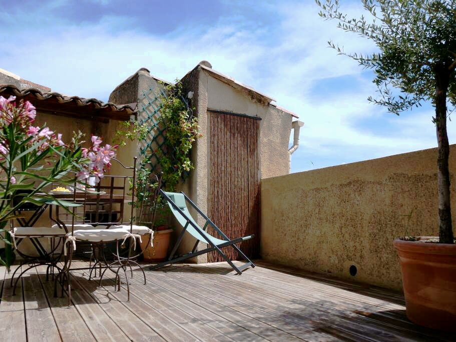 An outdoor terrace features a wooden deck, surrounded by potted plants and flowers. A table with chairs is placed beside a lounge chair, providing a space for relaxation. The terrace is enclosed by low walls, offering privacy and a view of the sky.