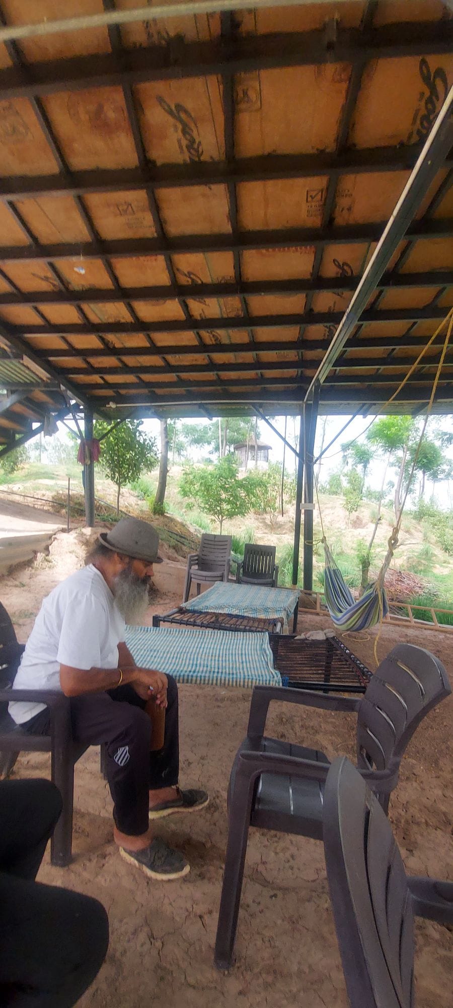 A shaded outdoor seating area features a long table surrounded by multiple chairs, situated under a rustic roof structure. Lush greenery and trees are visible in the background, contributing to a serene atmosphere, while a hammock is positioned nearby for relaxation.