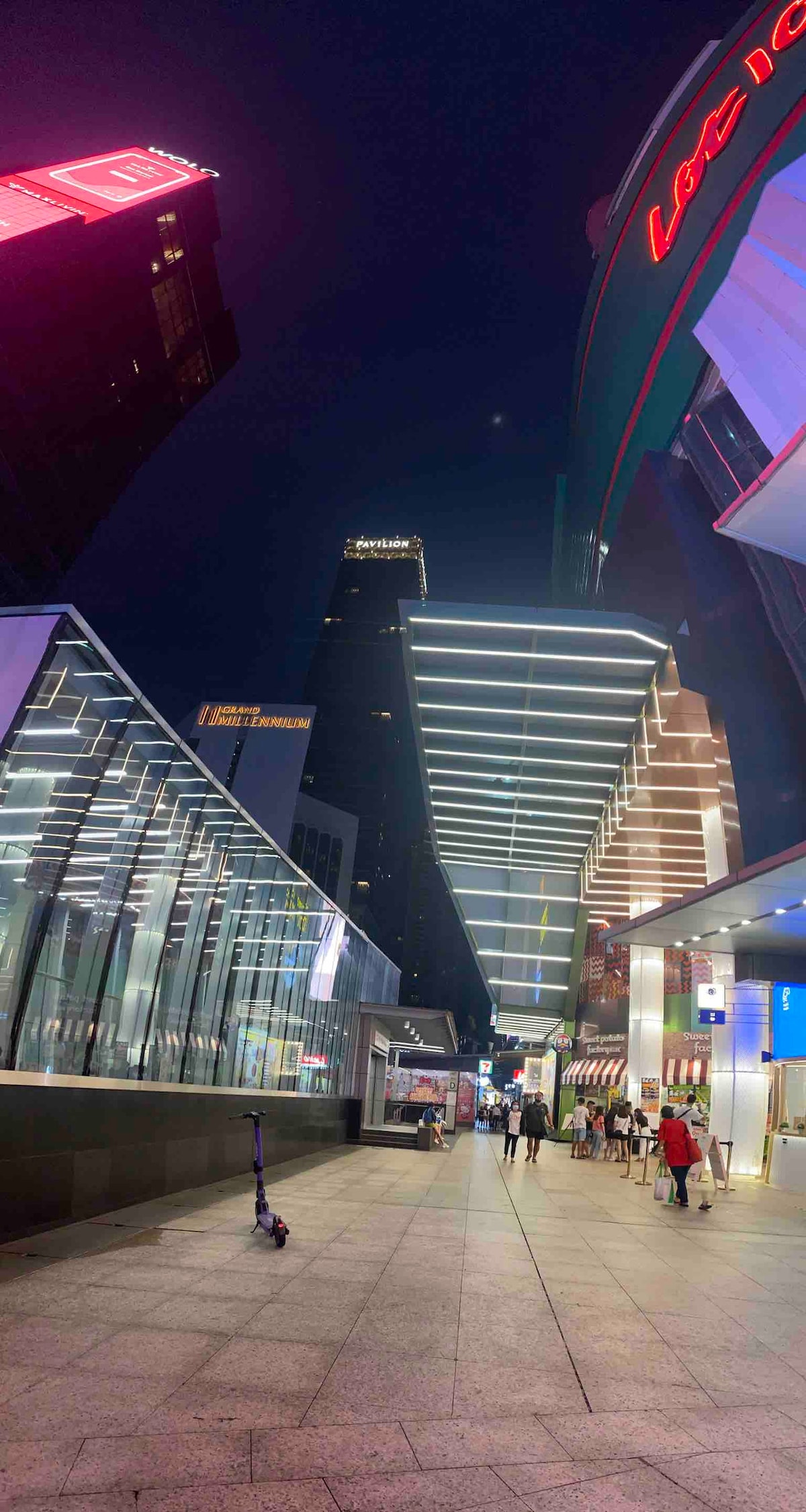 A vibrant city street is depicted at night, featuring modern architecture highlighted by colorful neon signage. The scene includes a wide pedestrian walkway, glass facades on either side, and distant skyscrapers reaching towards the sky.