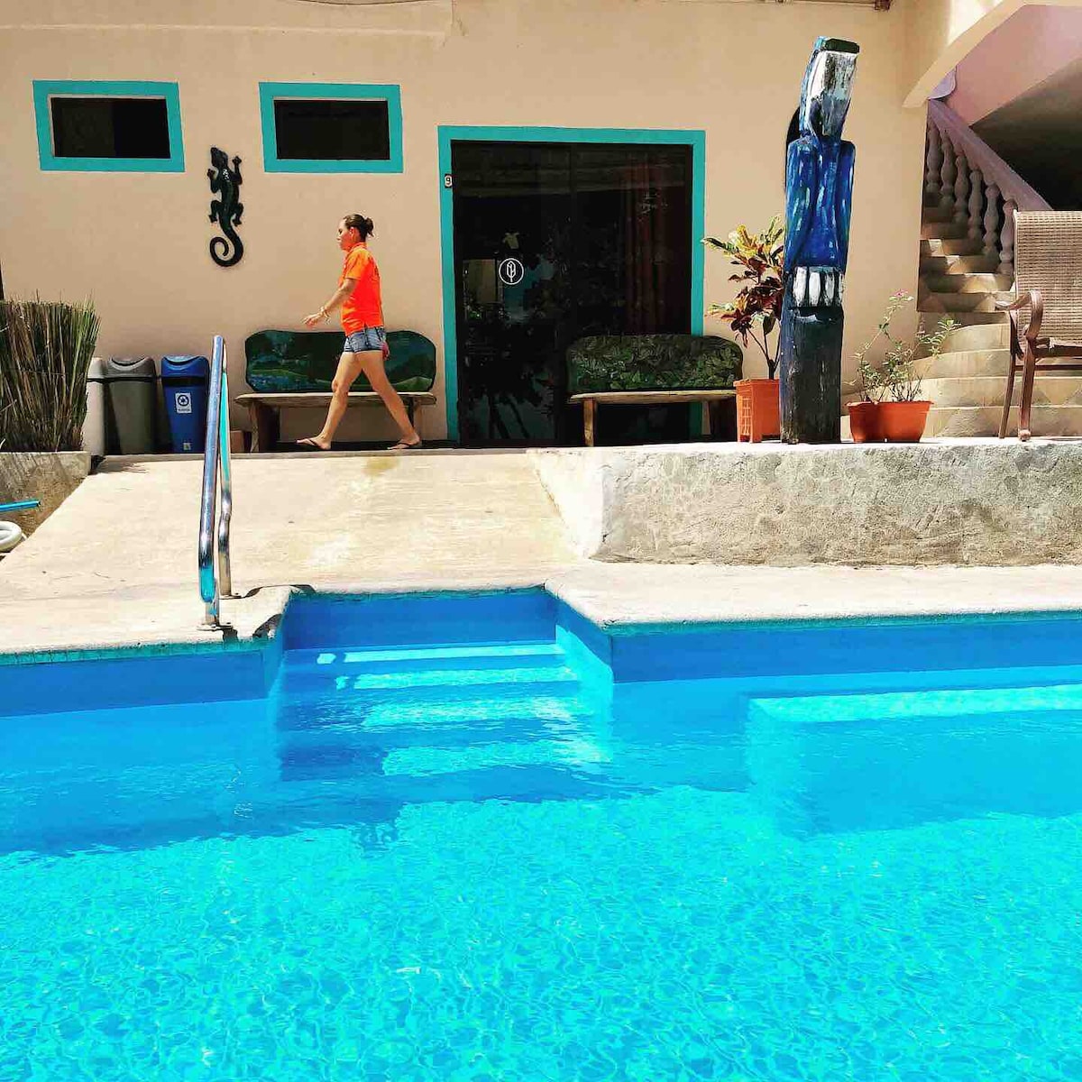 A clear blue swimming pool reflects the vibrant surroundings, with a person walking along the edge. The pool area is bordered by a building featuring a colorful facade, and a lounge area with seating is visible in the background, contributing to a relaxing atmosphere.