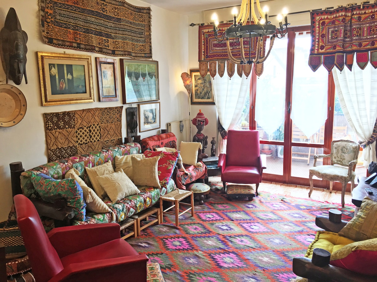 A vibrant living area features an eclectic mix of furnishings, including patterned sofas and two red armchairs. Colorful textiles adorn the walls, and artwork adds character. Large windows allow natural light to fill the space, with a warm, inviting ambiance created by a traditional chandelier.