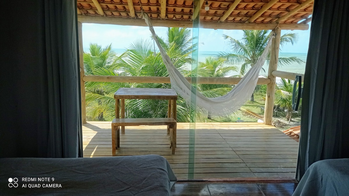 A balcony area offers views of the ocean and lush palm trees. A wooden table is set on the deck, complemented by a hanging hammock. Natural light filters through sheer curtains, enhancing the serene outdoor living space.