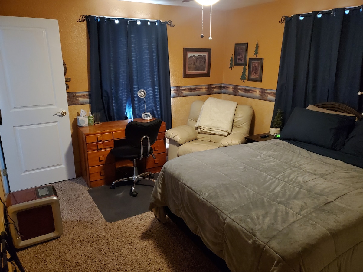 A cozy bedroom features a queen bed covered with a soft gray blanket. A desk with a comfortable chair is situated near the window, and a recliner is placed beside it. Light from the window is filtered by dark curtains, while warm wall colors create a comforting atmosphere.