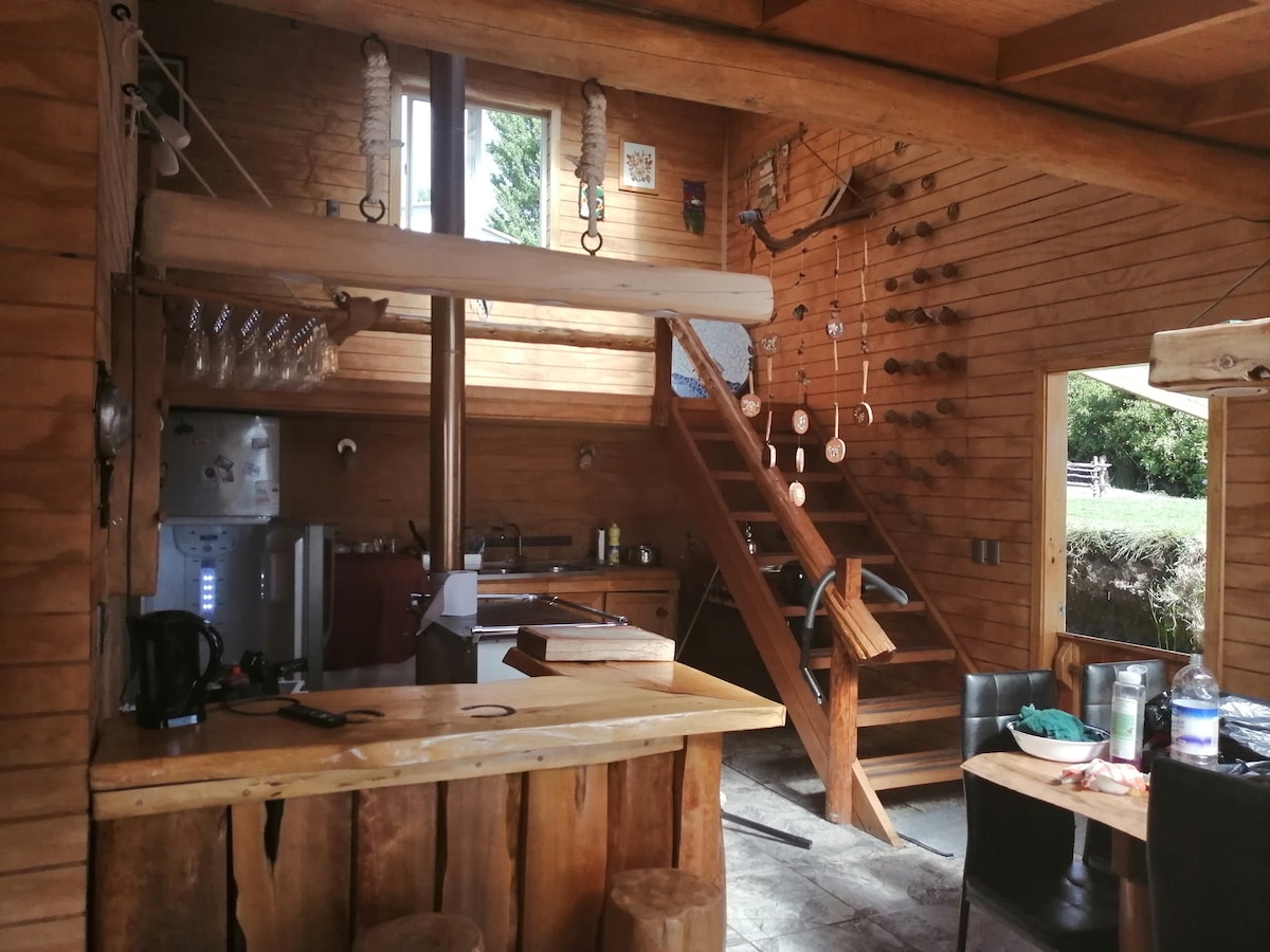 The interior features a rustic wooden design with a central staircase leading to the upper level. A well-equipped kitchen is visible alongside a bar area made of logs. Natural light fills the space through large windows, showcasing the inviting atmosphere.