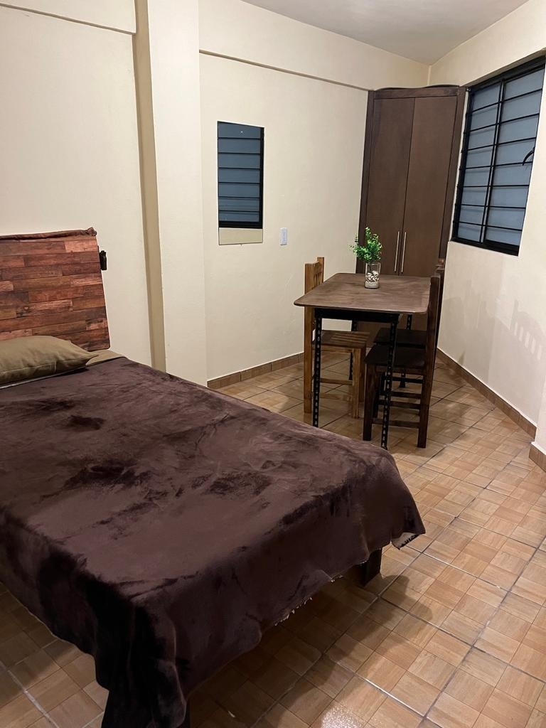 A cozy sleeping area features a single bed with a brown blanket and a small wooden table accompanied by two chairs. Natural light enters through the window, enhancing the room's serene ambiance. A wardrobe is present, providing additional storage space.