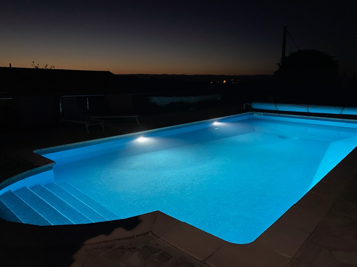 The illuminated pool, set against a night sky, features a soft glow reflecting off its water surface. Subtle light enhances the pool steps, inviting evening swims. The surrounding area is tranquil, offering a peaceful ambiance.