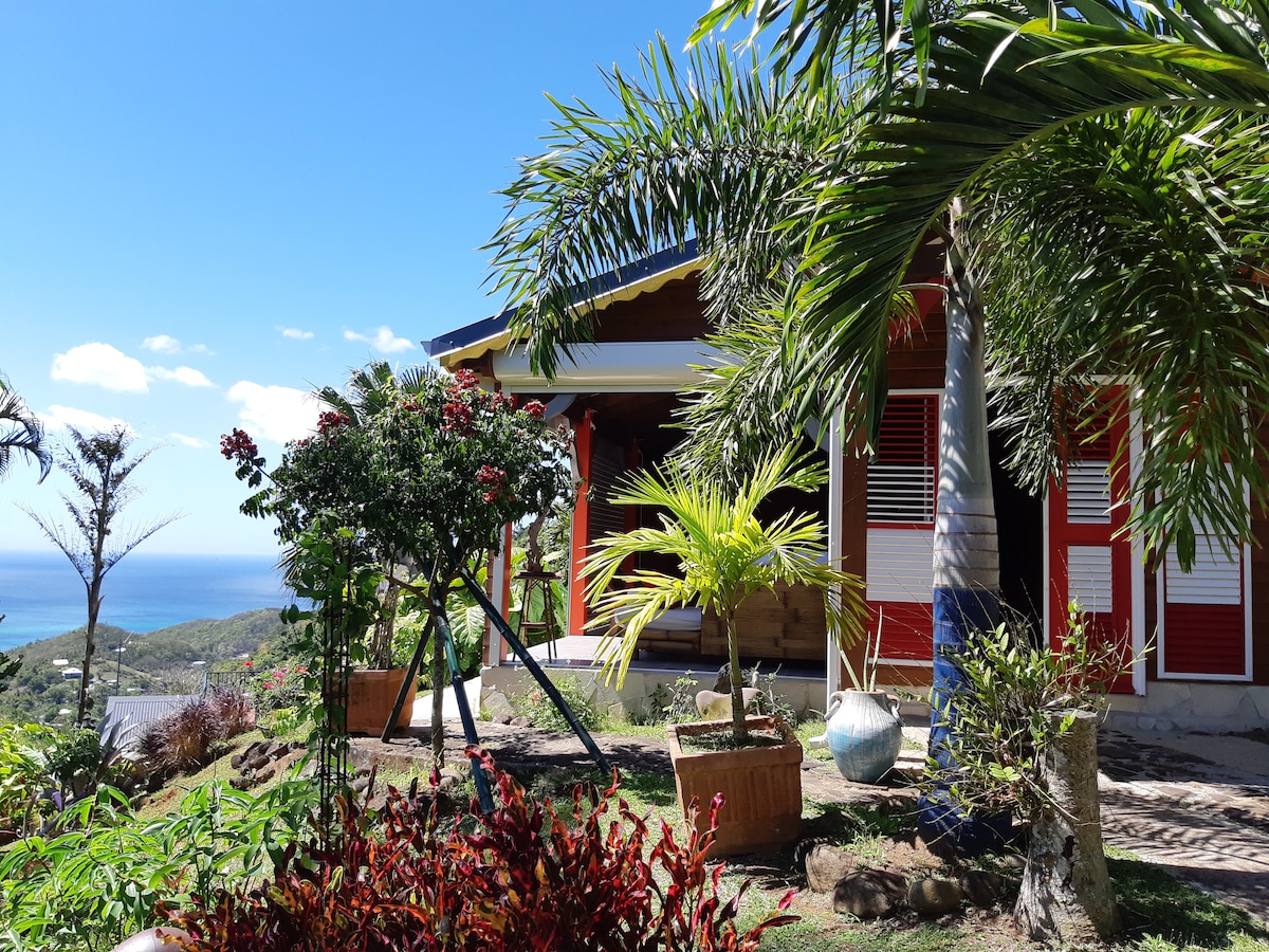 A vibrant garden surrounds the colorful Creole-style gîte, featuring a combination of tropical plants and flowers. The gîte's exterior is adorned with red shutters and large windows, providing unobstructed views of the Caribbean Sea and lush landscape beyond.