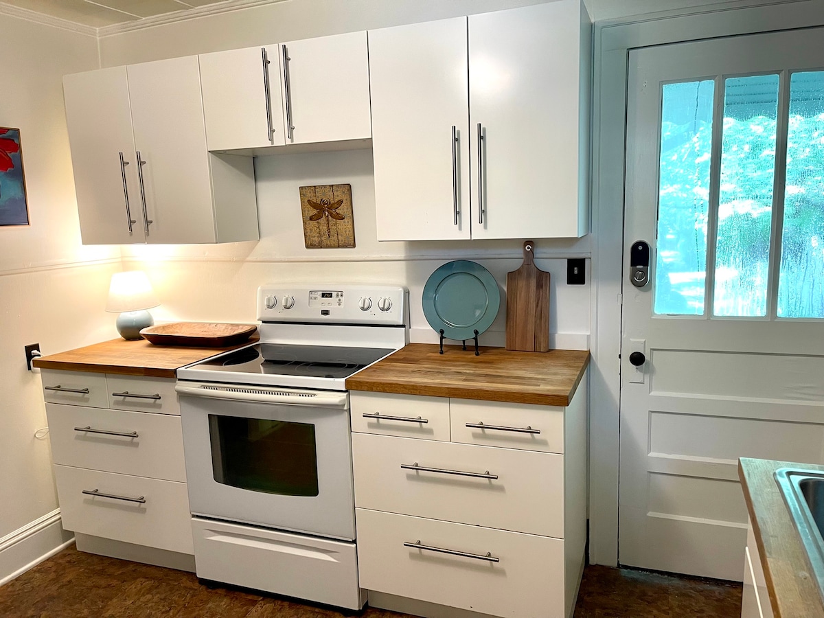 A modern kitchen features sleek white cabinetry and a wooden countertop. Standard appliances include an oven and a stove, while a soft glow is provided by a small lamp. A door with a window offers access to the outside, enhancing natural light in the space.