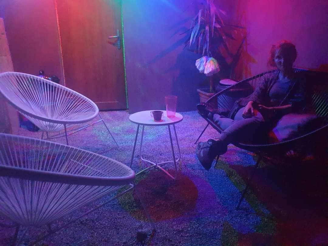 A cozy outdoor seating area is illuminated by colorful lights, featuring two white lounge chairs positioned around a small table. A person is seated comfortably, with a plant visible in the background, creating a relaxed environment for social gatherings.