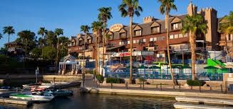 A multi-story building is seen along a waterfront, featuring a mix of balconies and outdoor seating areas. Palm trees line the property, contributing to a tropical ambiance. Multiple boats are docked along the dock, ready for use in the nearby water activities.