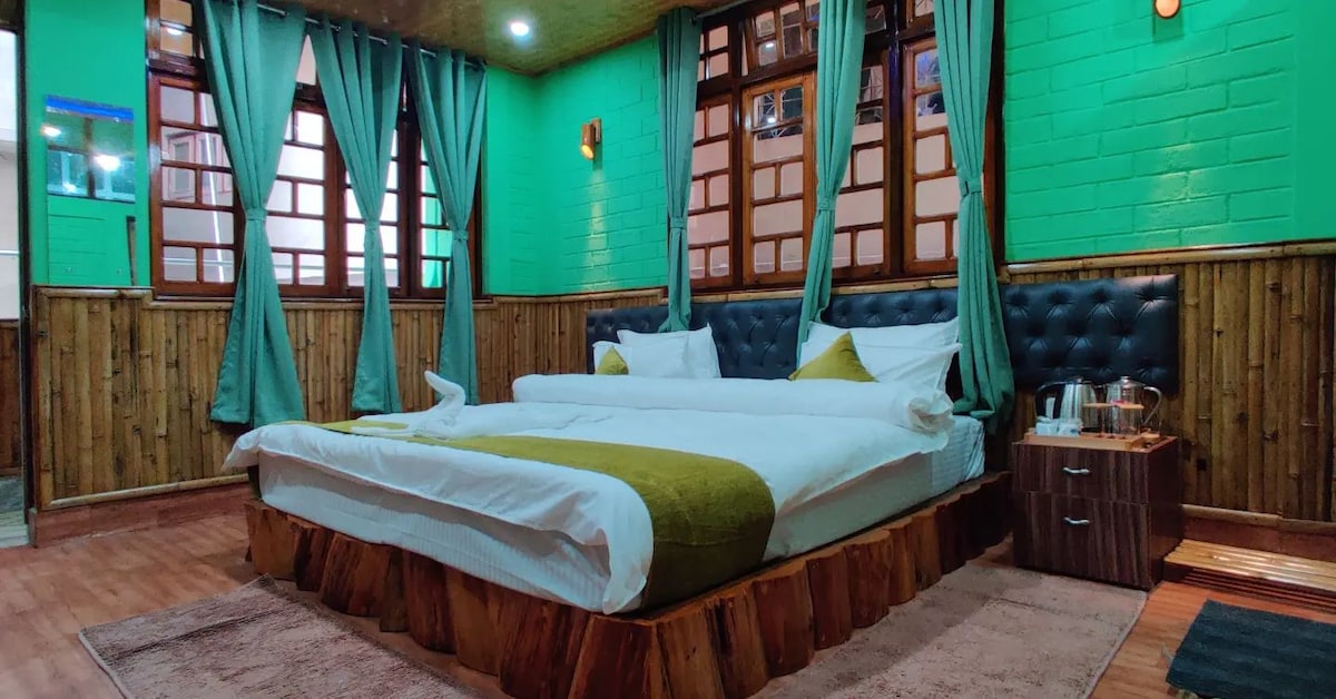 A spacious sleeping area features a large bed with a green and white color scheme. The walls are adorned with vibrant green paint and wooden paneling. Curtains frame the windows, and a bedside table holds a small lamp and glassware.