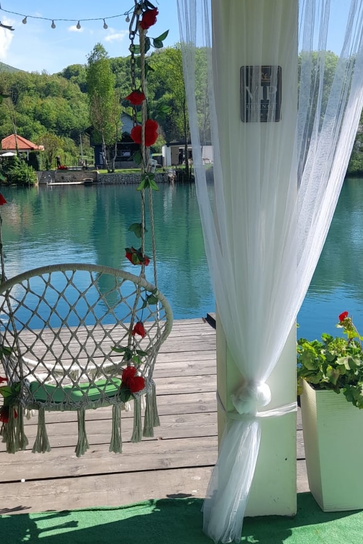 A hanging chair adorned with decorative flowers is positioned on a terrace, overlooking a serene body of water. Lush greenery surrounds the area, and gentle curtains sway with the breeze, inviting relaxation in the natural setting.