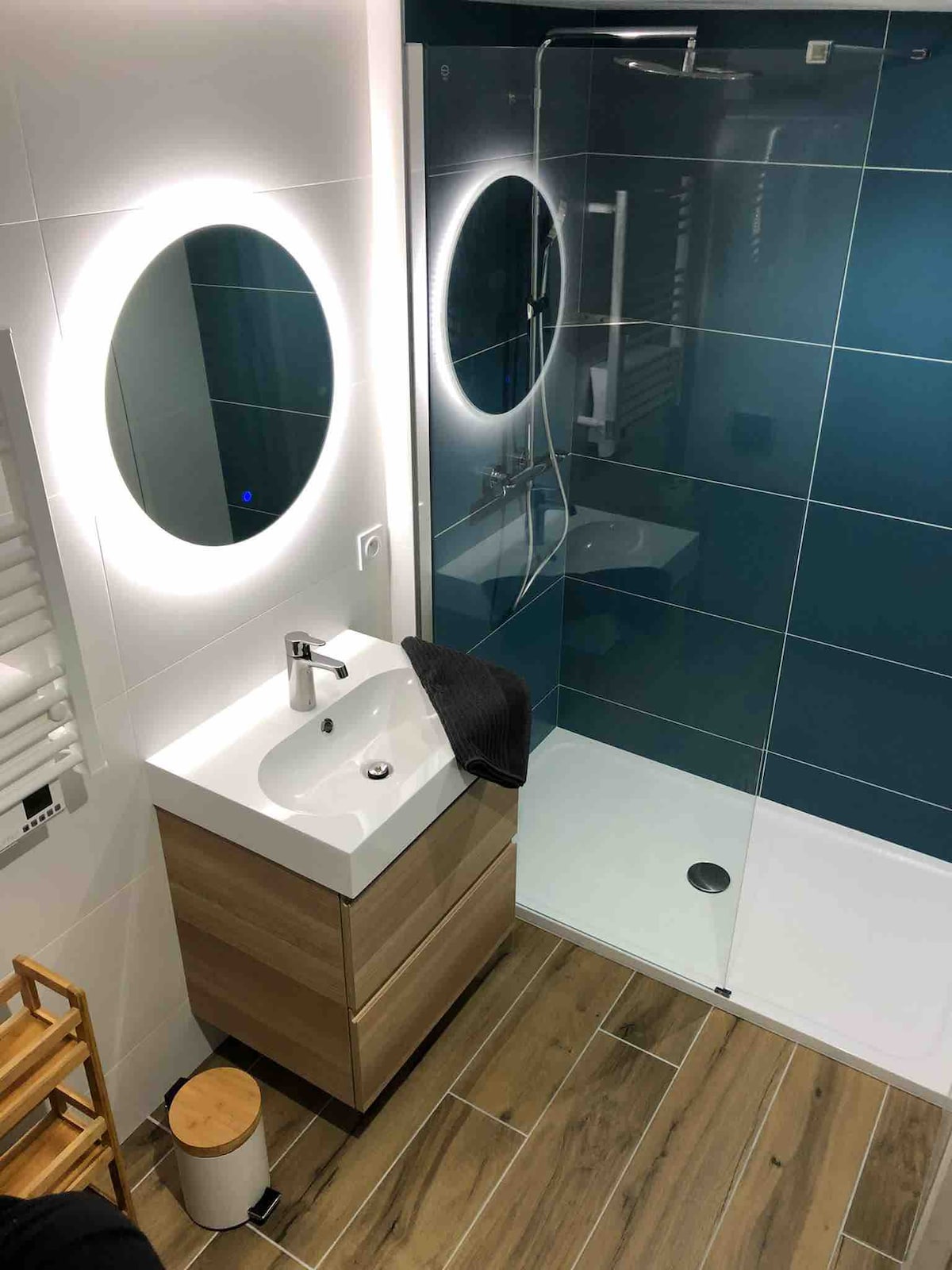 A modern bathroom features a glass shower with sleek fixtures and a vanity with a round mirror. The wall is accented with dark blue tiles, while wood-look flooring complements the neutral tones. Additional storage is provided by a small wooden stool and a towel rack.