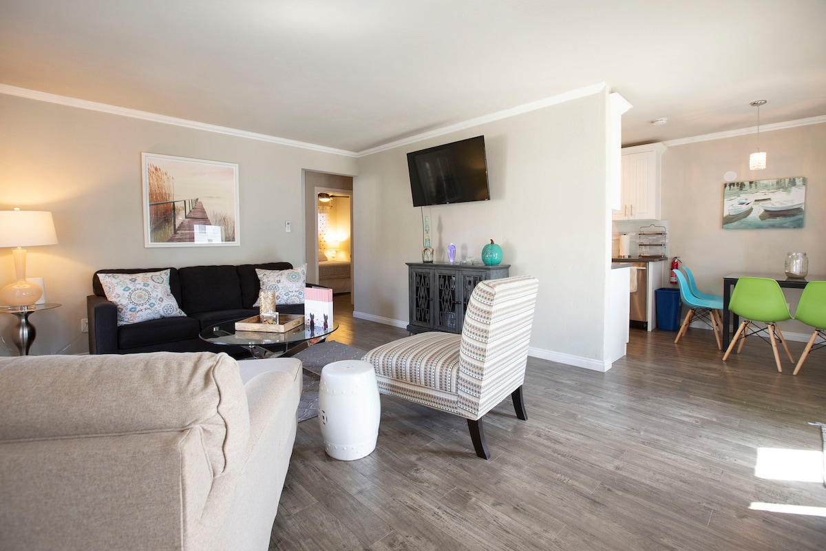 A modern living space features a black sofa with decorative pillows, complemented by a light-colored armchair. A sleek coffee table sits in front, and a wall-mounted television is visible. The open layout includes a dining area with colorful chairs, enhancing the room's inviting feel.