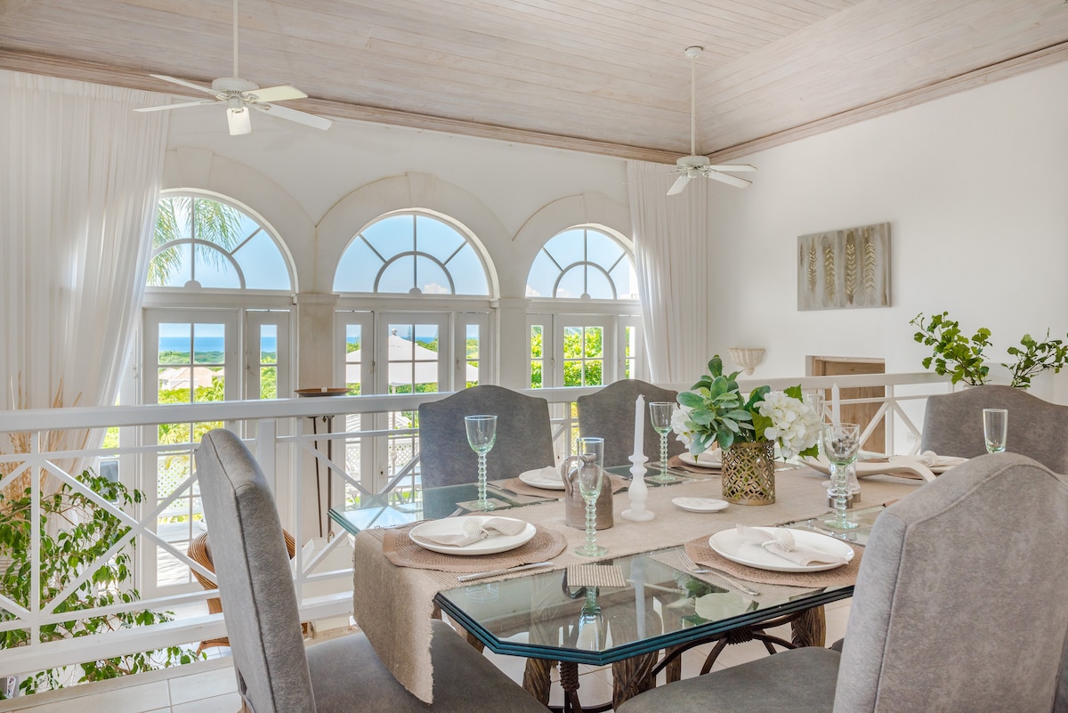 A dining area is presented with a glass table surrounded by plush gray chairs. Natural light filters through large arched windows, offering views of the ocean. Decorative elements include elegant tableware and a floral arrangement, enhancing the serene atmosphere of the space.