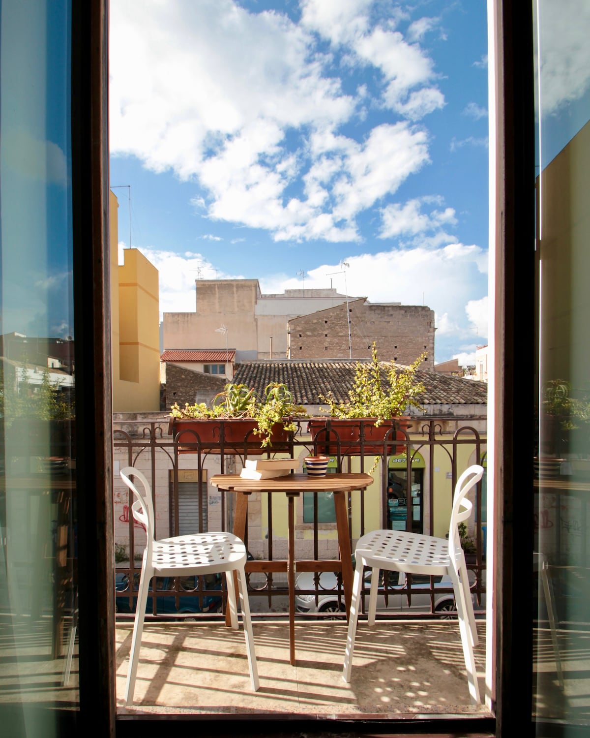 A balcony view is presented, featuring a small table surrounded by two white chairs. Potted plants add a touch of greenery, while a clear blue sky with scattered clouds enhances the lively atmosphere of the space.