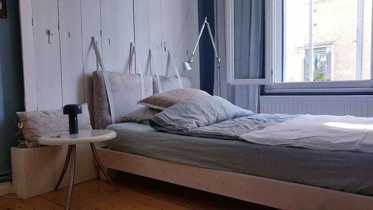 A bright bedroom features a spacious bed with soft linens and an assortment of pillows. A small round table is positioned beside the bed, and a lamp provides focused light. Natural light enters through the window, which is dressed with light curtains, illuminating the wooden floor.
