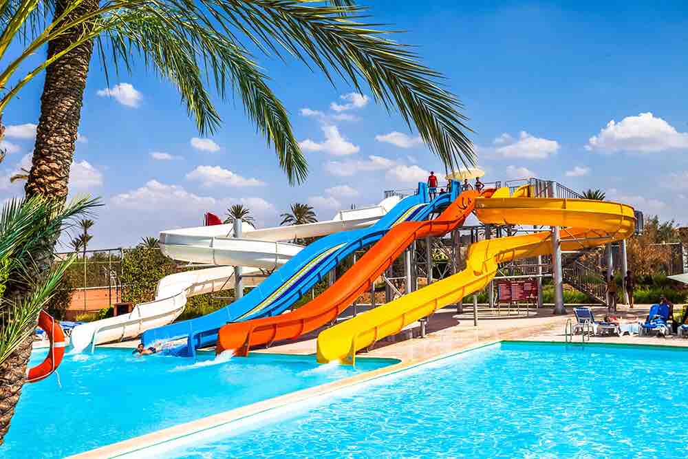A vibrant water park features multiple colorful water slides in shades of red, blue, yellow, and white, positioned alongside a sparkling pool. Lush palm trees are visible in the background, providing a tropical feel to the recreational area.