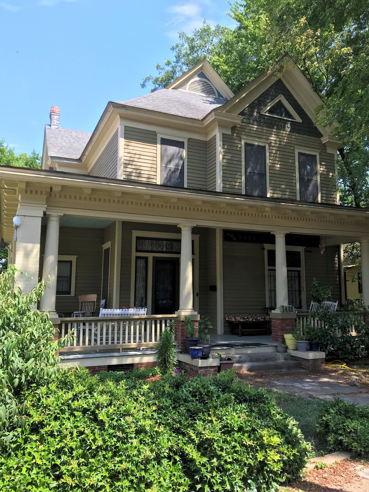 The exterior of a historic two-story house is shown, featuring a green and beige color palette. A welcoming front porch is lined with wooden chairs, surrounded by well-kept shrubbery and potted plants. Tall trees provide shade, contributing to a peaceful atmosphere.