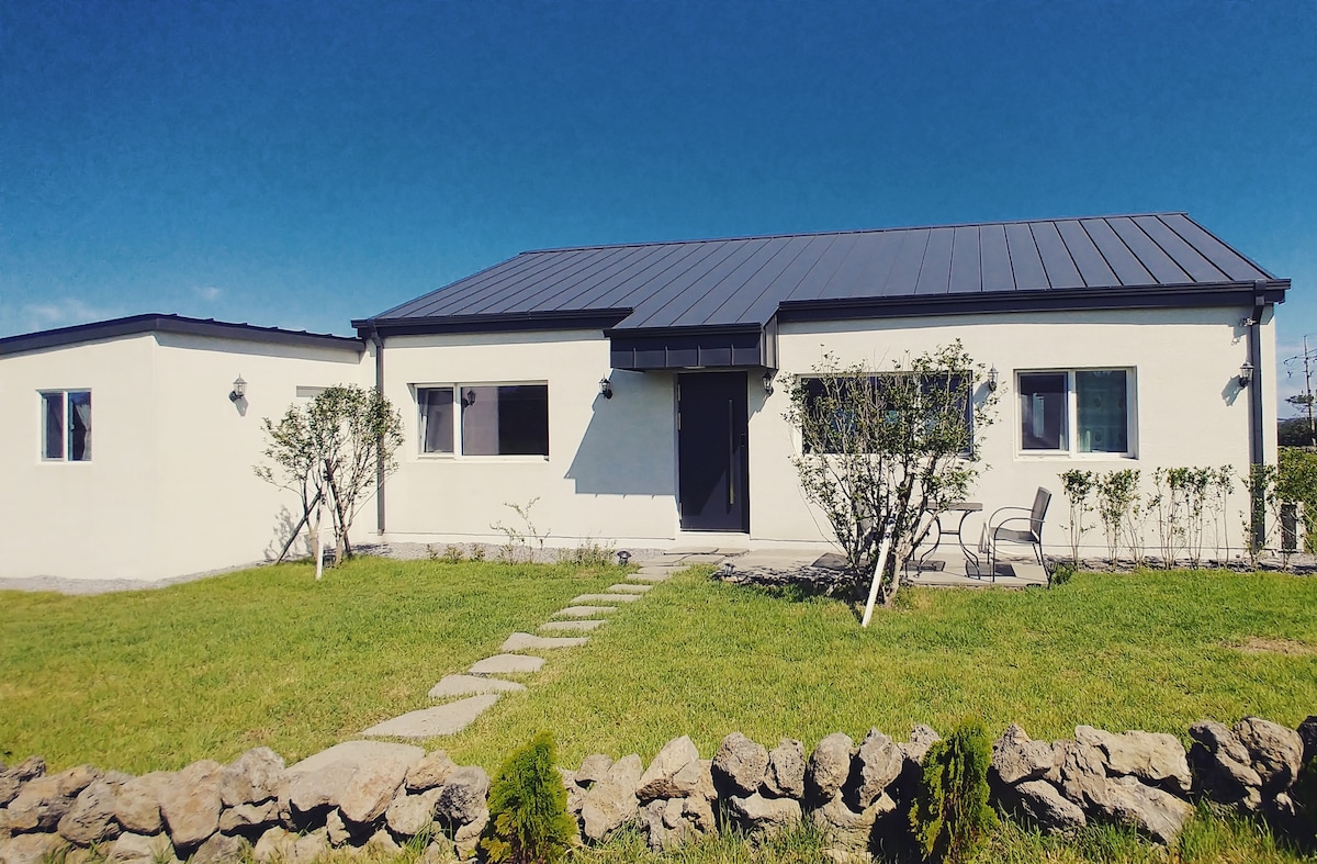 A single-story white building with a modern design is set against a bright blue sky. A well-maintained green lawn surrounds the property, featuring a pathway of flat stones leading up to the entrance. Two chairs are placed outside, framed by small shrubs.