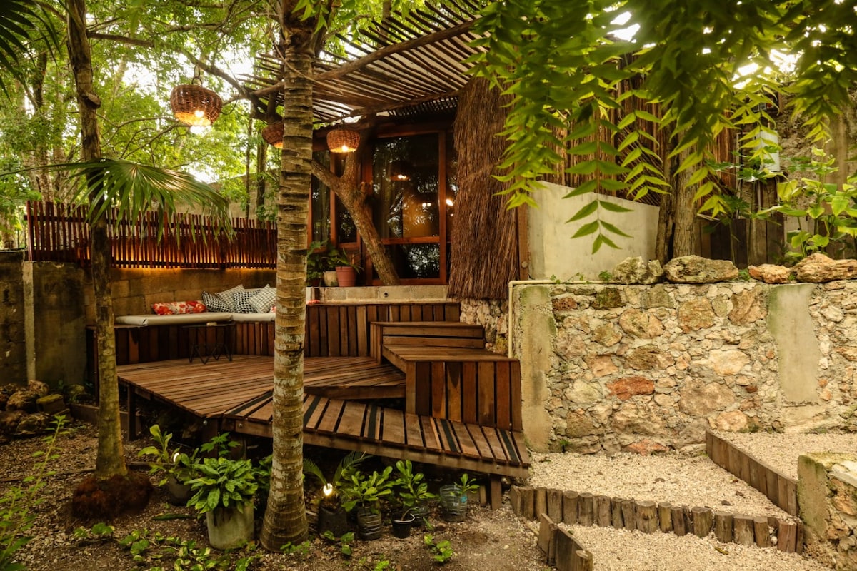 A secluded outdoor space is surrounded by lush greenery. A wooden deck features tiered steps leading to an inviting seating area with soft cushions. Natural elements are combined with thoughtful landscaping, complemented by warm lighting that enhances the tranquil atmosphere.