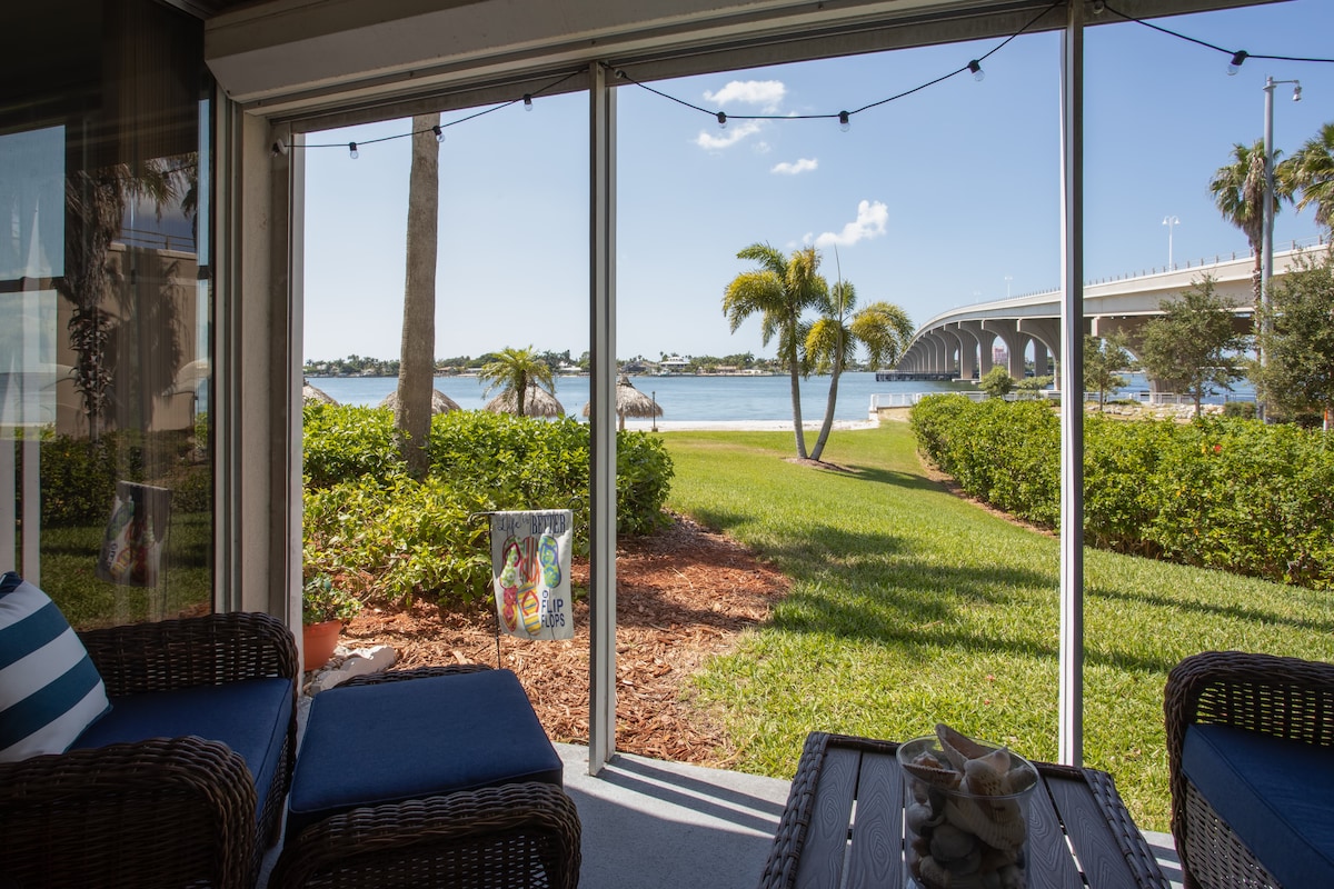 A cozy sitting area features comfortable wicker chairs and a small table, facing an expansive view of the serene water. Lush greenery and palm trees surround the space, while a bridge is visible in the distance under a clear blue sky.