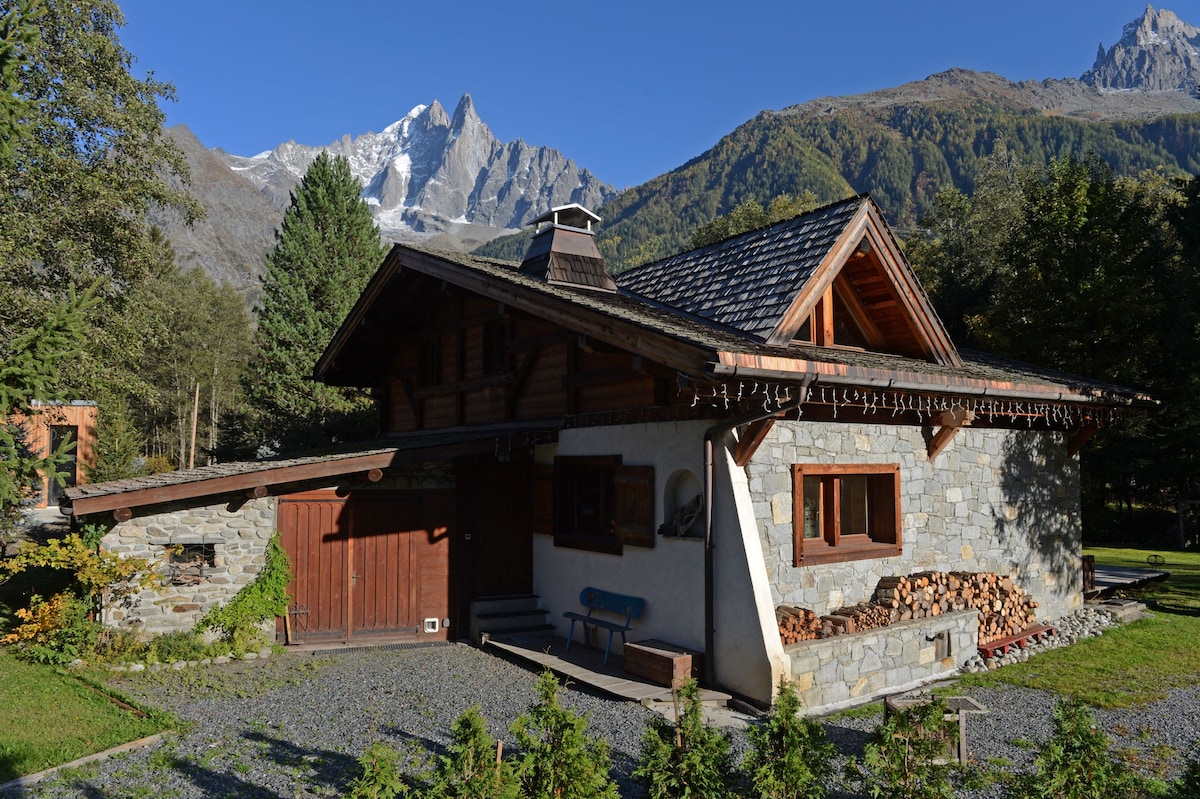 The chalet features a blend of stone and wood, set against a backdrop of majestic mountains. A sloped roof and decorative eaves reflect traditional alpine architecture. A wood pile is neatly stacked beside the entrance, while a gravel pathway leads to the front door, surrounded by lush greenery.