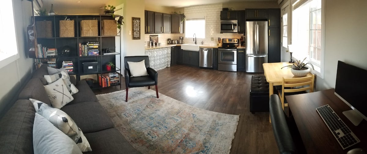 An open concept living area showcases a comfortable sectional sofa with patterned pillows. A dining area with a light wood table is visible, along with a modern kitchen featuring dark cabinetry and stainless steel appliances. Natural light enters through multiple windows.