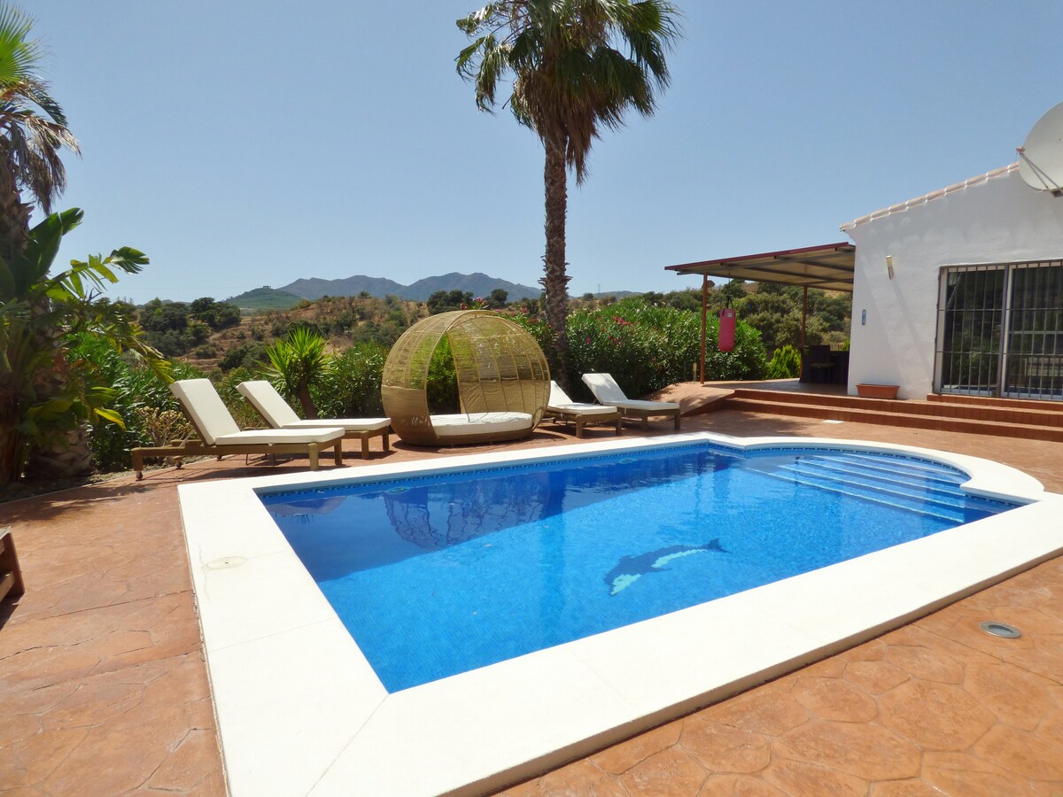 A private pool is framed by sun loungers under the shade of palm trees, surrounded by vibrant greenery. The clear water of the pool reflects the bright blue sky, with mountains visible in the background. A cozy seating area is partially shaded to the right.