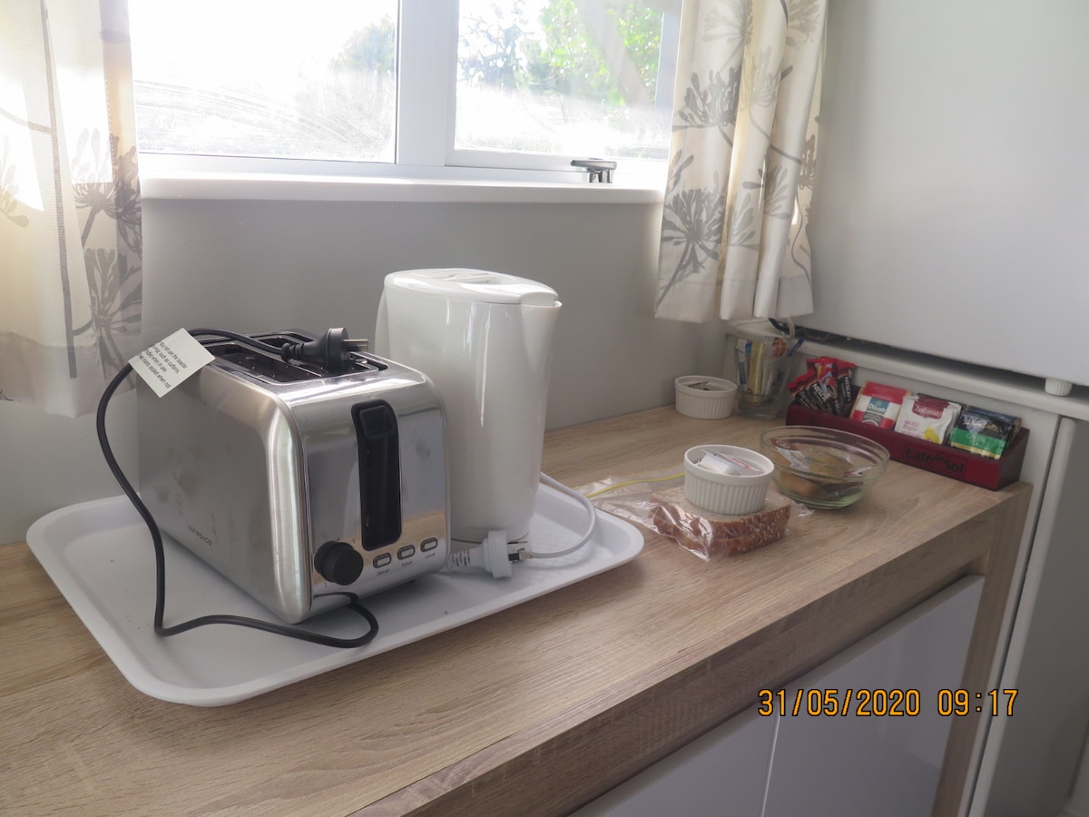 A kitchen countertop is shown, featuring a silver toaster and a white kettle positioned on a tray. Nearby, a small dish is filled with snacks, and various condiments are neatly organized in a red container. Natural light filters through the window adorned with floral curtains.