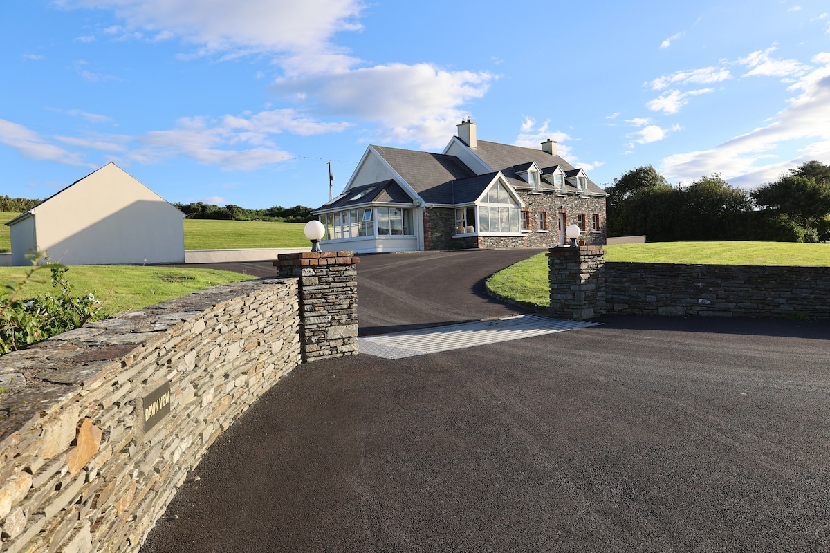 A spacious country home is set against a bright blue sky, flanked by lush green grass. The stone walls and gentle curves of the driveway lead toward the entrance, creating an inviting approach to the property.