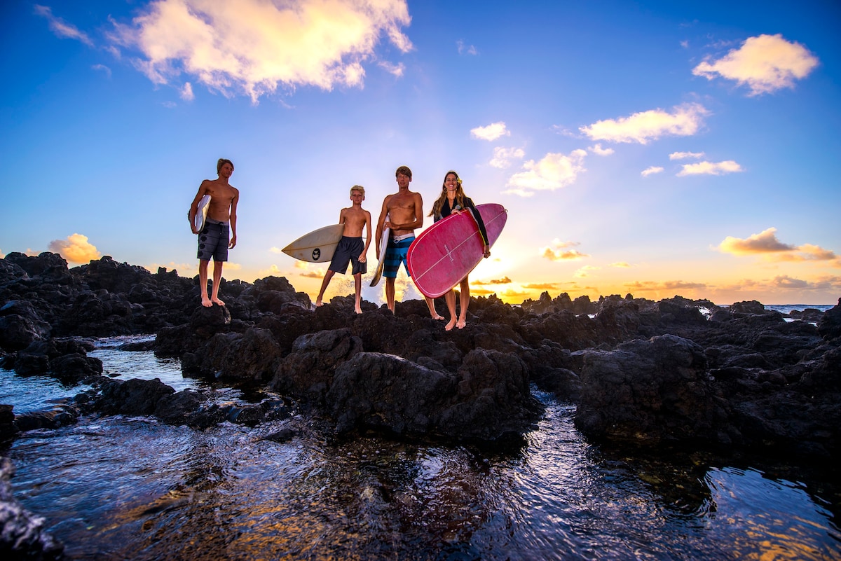 Four individuals stand on rocky coastal formations at sunset, holding surfboards. The vibrant sky features hues of orange, pink, and purple, reflecting on the water. The scene captures a serene yet lively atmosphere typical of Hawaii's surf culture.
