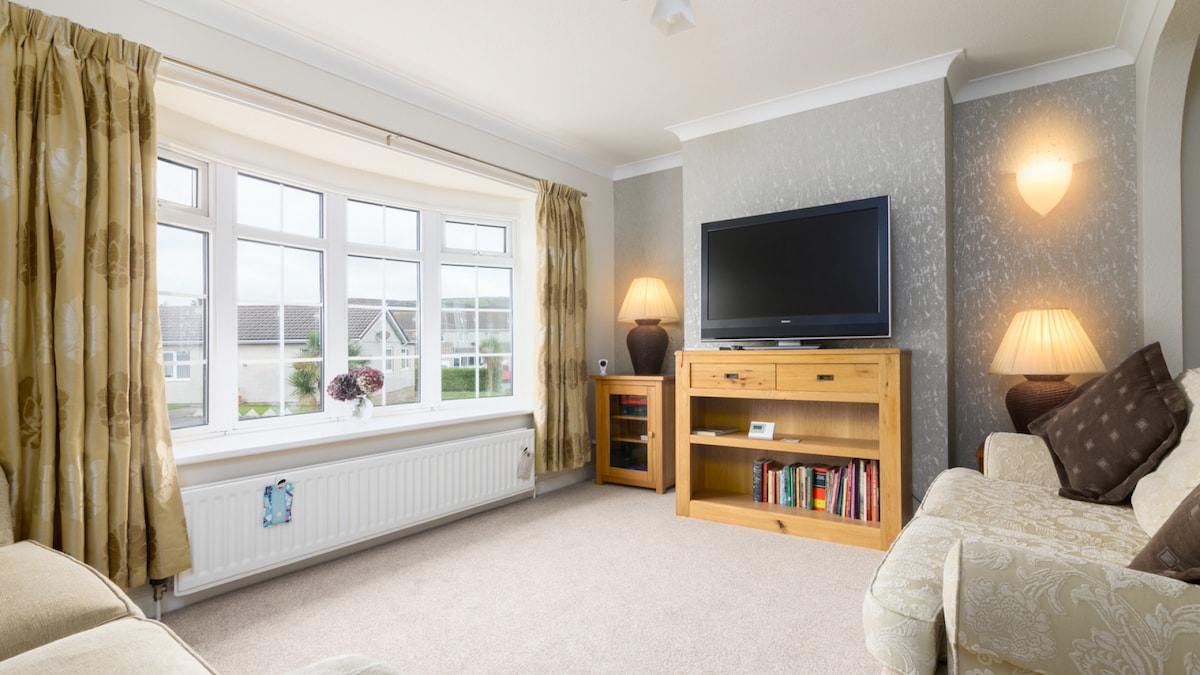 A well-lit lounge area features a large bay window with multiple panes, offering garden views. A wooden television unit with books is positioned beneath a flat-screen TV. Soft lighting from table lamps complements the warm-colored curtains and neutral-toned furnishings, creating a welcoming space.