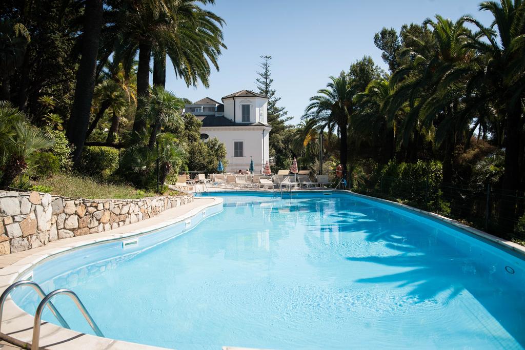 An outdoor swimming pool is surrounded by lush palm trees and well-maintained gardens. Sun loungers are positioned along the poolside, providing ample space for relaxation. A two-story building is visible in the background, offering a peaceful ambiance.