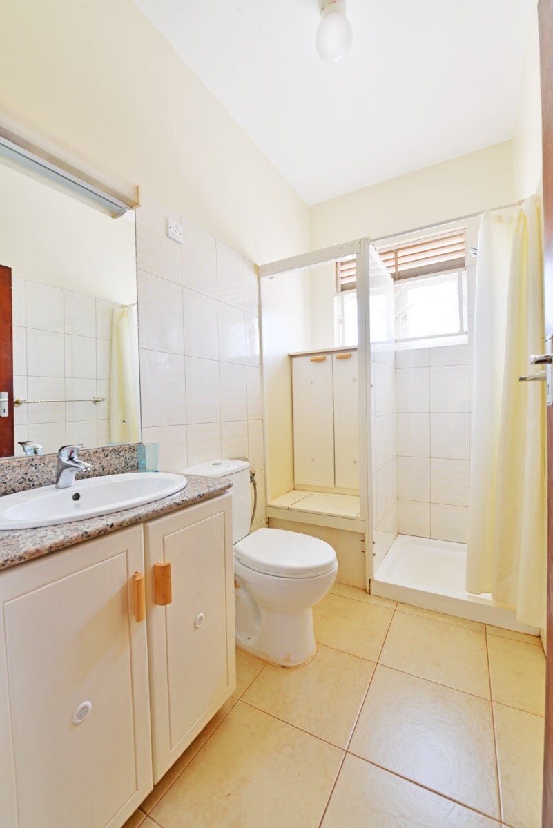 The bathroom features a clean design with a granite countertop and a mirror above the sink. A standing shower is enclosed with clear glass, while natural light enters through a window partially covered by a curtain. Neutral tones dominate the tiled space.