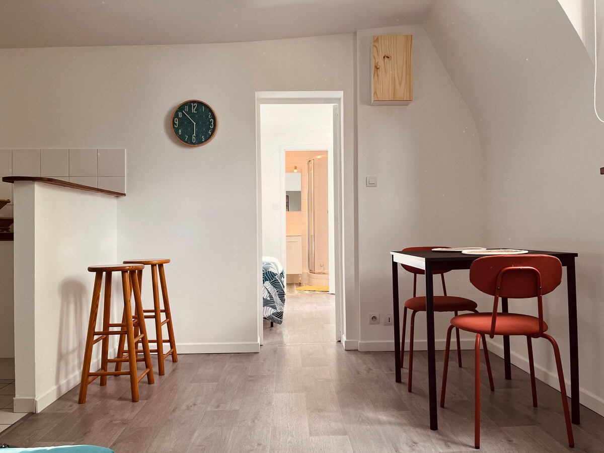 An open living space features a kitchenette with bar stools and a dining area. A small round table with two chairs is positioned beside the entrance. Light wood flooring and neutral wall colors create a bright ambiance, while a clock hangs on the wall, adding a functional detail.