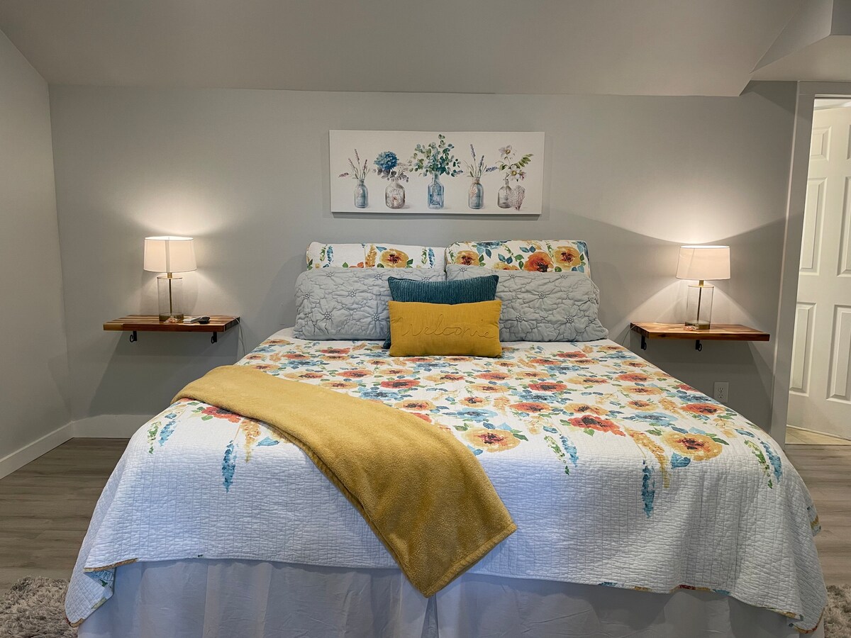 A spacious bedroom is highlighted by a king-sized bed adorned with floral bedding and accent pillows. Two bedside tables with lamps are positioned on either side. A decorative wall art piece featuring vases is displayed above the bed, providing a calm atmosphere.