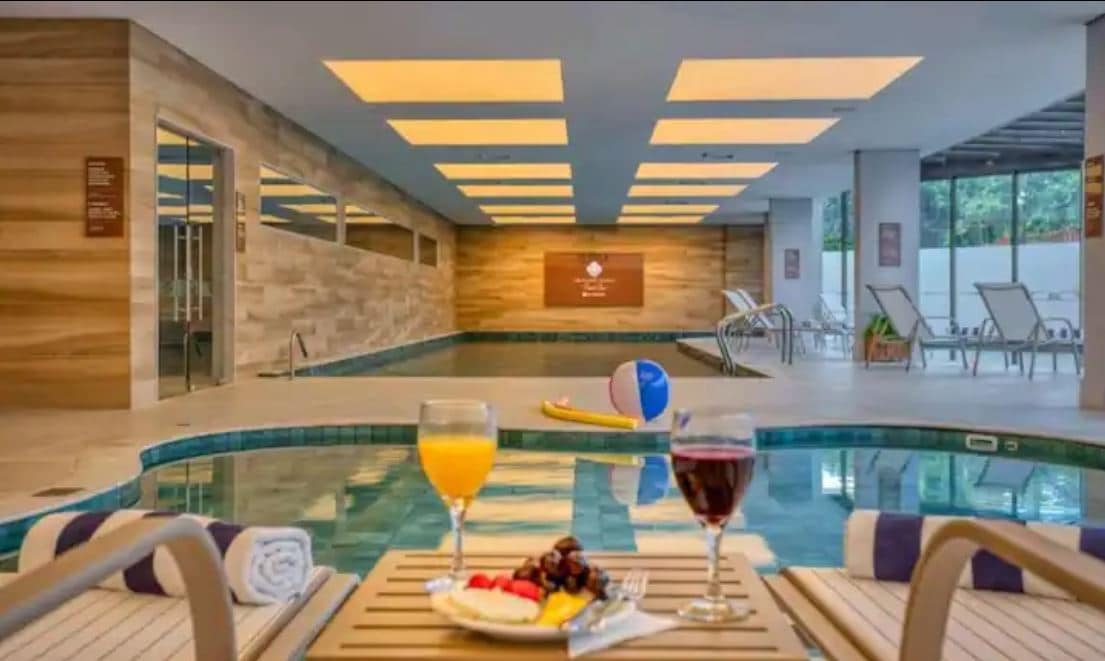An indoor swimming pool is visible, featuring a warm ambiance created by soft lighting. In the foreground, a table holds two glasses of drinks and a plate of colorful fruit, accompanied by neatly rolled towels. Lounge chairs are positioned along the pool's edge.