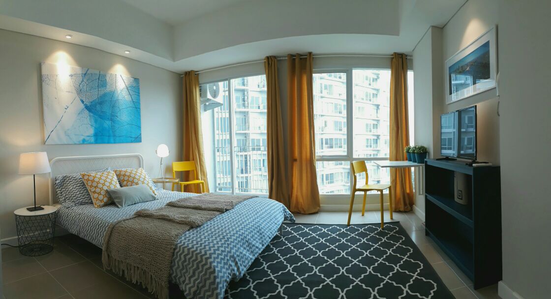 A modern bedroom features a spacious bed covered with textured bedding. A large window provides natural light, framed by yellow curtains, with a city view visible outside. A small dining table and two yellow chairs offer additional seating, complemented by a stylish area rug.