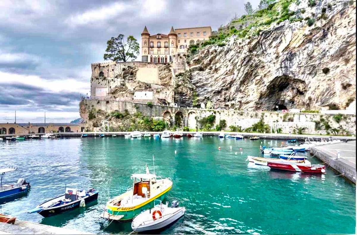 A harbor scene showcases an array of small boats anchored in calm turquoise waters. A striking cliffside building rises above the marina, with lush greenery and rocky outcrops framing the background, while cloudy skies create a dramatic atmosphere.