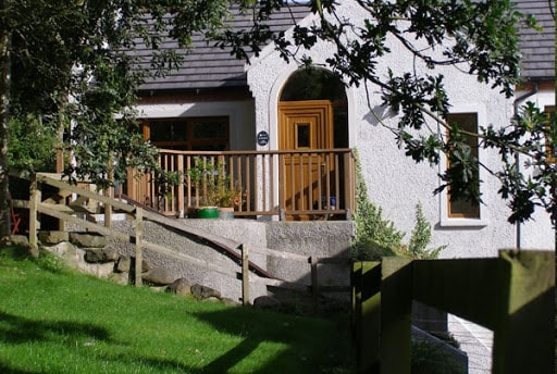 The entrance to the cottage is marked by a wooden door with a decorative archway. Flanked by a handrail, a gently sloping pathway leads from the lawn to the entrance. The surrounding area features greenery and small shrubs, enhancing the cottage's natural setting.