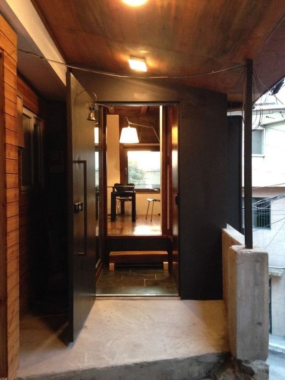 An entrance is framed by dark wooden panels and a sleek black door, which stands open to reveal a well-lit interior space. A wooden bench is positioned just inside, while minimalistic furniture is visible beyond, indicating a modern and inviting atmosphere.