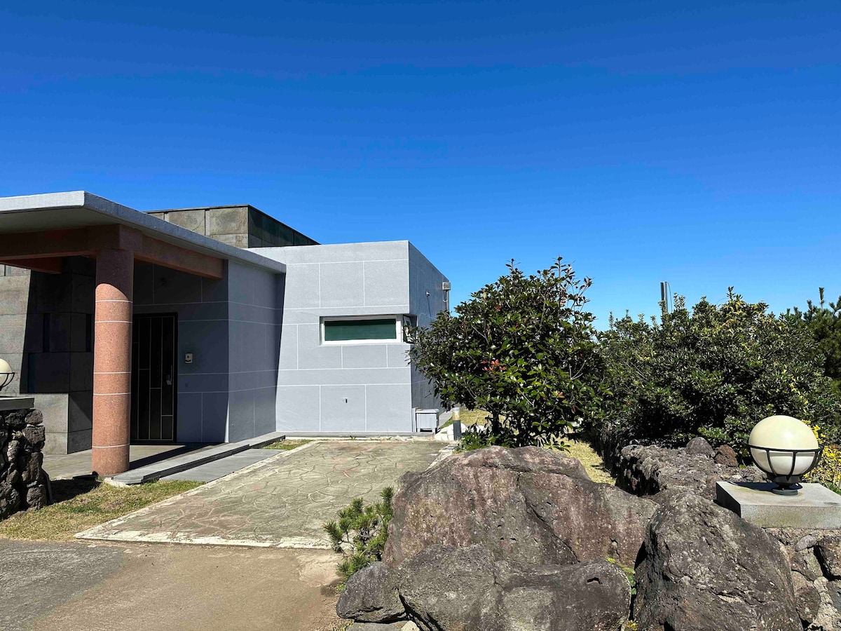 A modern single-story home with a light grey exterior is surrounded by greenery. A stone pathway leads to the entrance, framed by neatly trimmed bushes and rock features. The clear blue sky provides a bright backdrop, enhancing the peaceful setting.